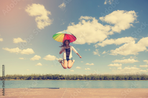 young woman jumping with holding colorful umbrella beside river