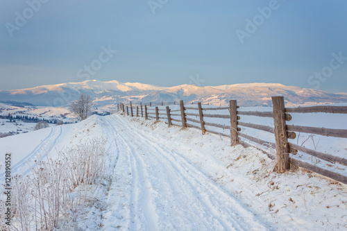 Winter country landscape with timber fence and snowy road into evergreen forest