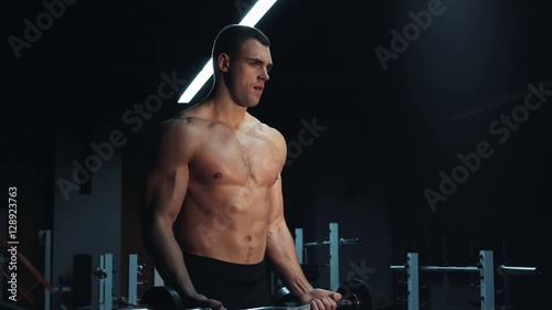 Muscular man doing crossfit training in a dark shadowy gym lifting weights holding a barbell at waist level in a health and fitness concept, movement round close shot photo