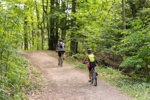 Couple of people cycling together in the woods down a dirt path on a beautiful summer day. Green canopy of trees above them. View from the back of people - full body.