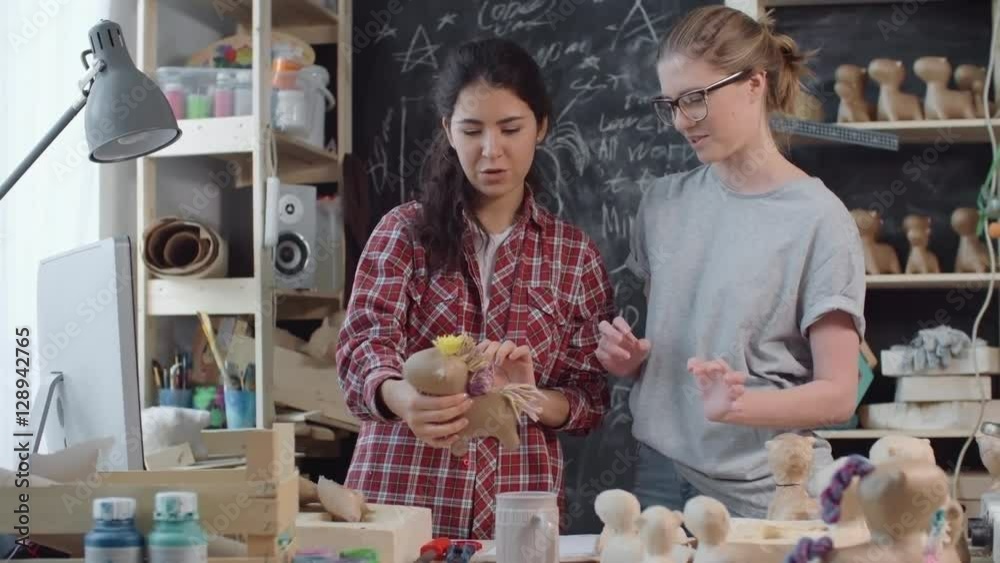 Happy young women standing in workshop and looking at finished papier-mache horse toy, then celebrating and hugging 