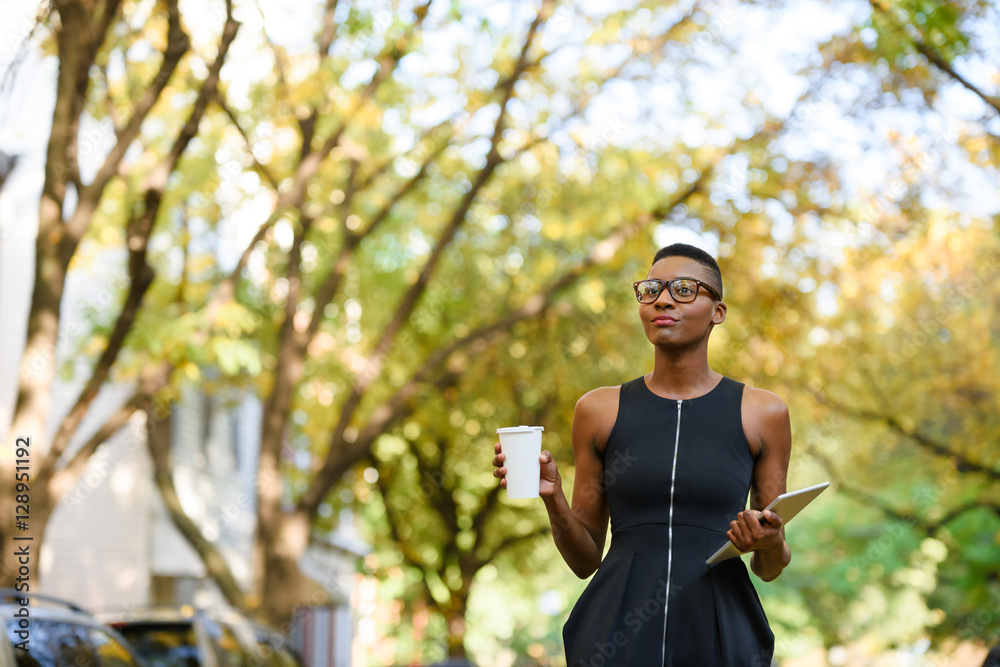 Obraz premium Fashionable businesswoman walking outside with tablet and coffee cup wearing glasses.