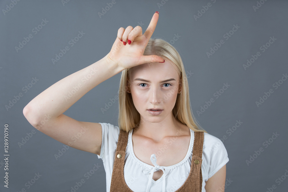 School blonde girl showing loser sign studio gray background School blonde girl showing loser sign studio gray background