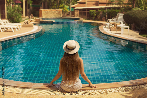 Young woman with long hair in hat sitting near the swimming pool, enjoy summer and holiday © Annatamila