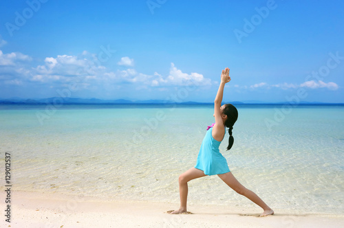 Young healthy Asian kid doing yoga on tropical beach in warrior pose I. Preteen girl in swimwear practice yoga on sea beach blue sky sand sun daylight relaxation landscape for sun protection ad.
