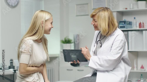 Mid Adult Female Doctor Consults Young Female by Showing to Her Tablet Computer with X-Ray. Woman Hodls her Stomach. Both Smile. Shot on RED Cinema Camera in 4K (UHD). photo