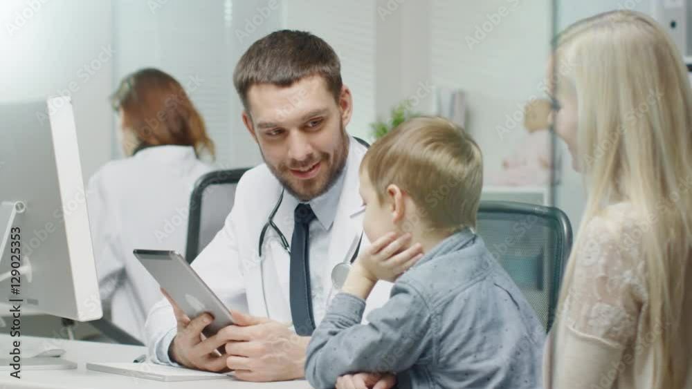 Male Doctor Consults Young Boy and His Mother with a Help of a Tablet. They Smile and Joke Warmly. Shot on RED Cinema Camera in 4K (UHD). Male Doctor Consults Young Boy and His Mother with a Help of a Tablet. They Smile and Joke Warmly. Shot on RED Cinema Camera in 4K (UHD).