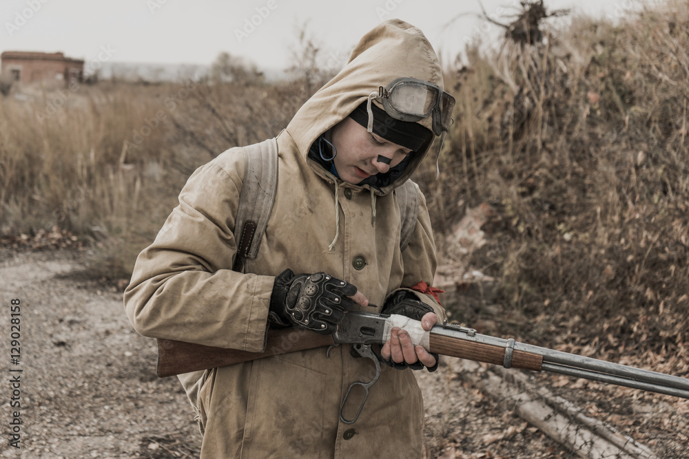 Fototapeta premium wandering boy. boy with a gun. boy goes to an abandoned building. boy stands in front of a building. Post apocalypse. Boy traveling on foot in a post-apocalyptic world in search of food. 