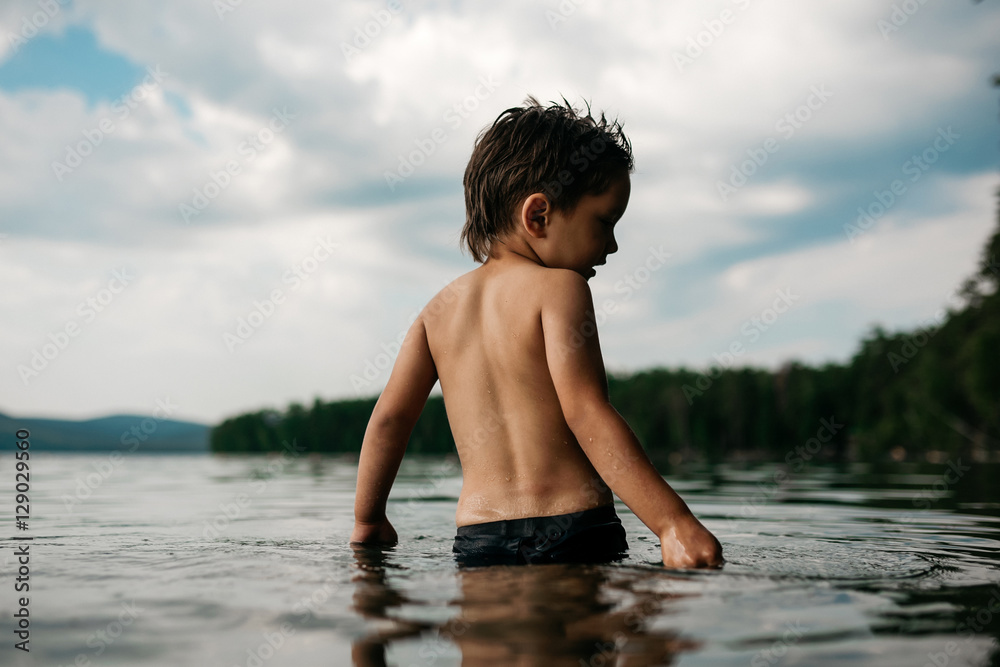 Little boy 4 years is waist-deep in the water of forest lake. View from the back. Beautiful nature, water surface, forest and mountains on the horizon Little boy 4 years is waist-deep in the water of forest lake. View from the back. Beautiful nature, water surface, forest and mountains on the horizon