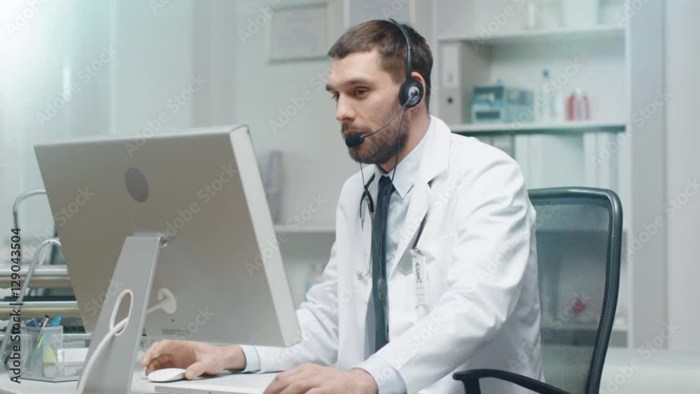 Male Doctor Consults Patients on the Internet. He Sits before His Personal Computer and Wears Microphone. Shot on RED Cinema Camera in 4K (UHD). Male Doctor Consults Patients on the Internet. He Sits before His Personal Computer and Wears Microphone. Shot on RED Cinema Camera in 4K (UHD).