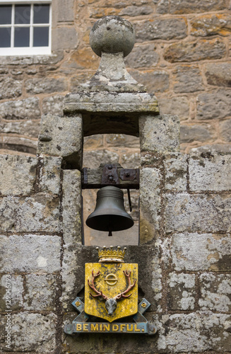 Cowdor, Scotland - June 2, 2012: The black bell hangs in gray stone niche. Golden coat of arms and slogan, Be Mindful. In the arch above the entrance gate of historic Cowdor Castle. 