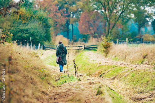 Woman with shoulder bag walking in field with autumn trees.