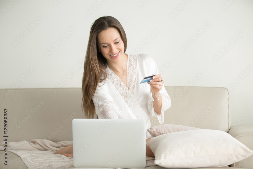 Young smiling woman wearing a dressing gown shopping online, using credit card for e-commerce transactions, making donation, checking personal finance Young smiling woman wearing a dressing gown shopping online, using credit card for e-commerce transactions, making donation, checking personal finance