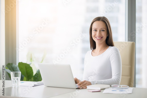 Young smiling female receptionist at the modern office desk with a laptop ready to greet clients, customers and visitors, direct them, free consultations, first impression. Looking at the camera