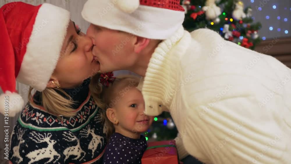 Young father gives a Christmas gift to a little daughter and kisses wife. Medium shot Young father gives a Christmas gift to a little daughter and kisses wife. Medium shot
