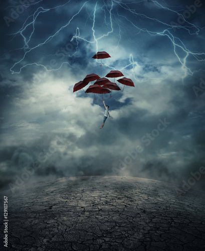 Young man with a lot of umbrellas as an air balloon rise up to the sky full of lightnings, above the cracked desert ground. Risk and success concept. photo