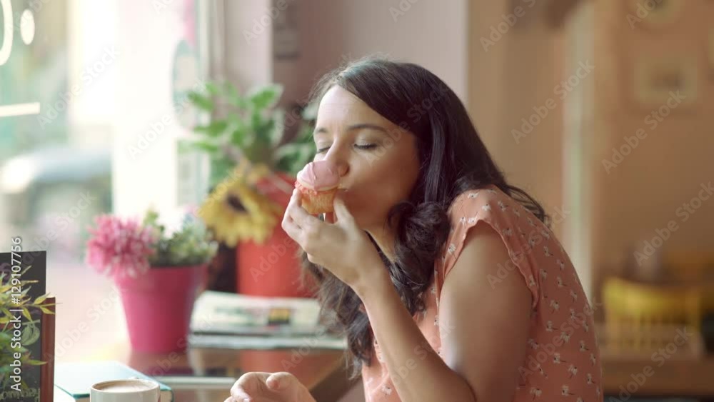 Young woman eating a cupcake next to the window in a coffee shop. Funny moment. She smiles. Young woman eating a cupcake next to the window in a coffee shop. Funny moment. She smiles.