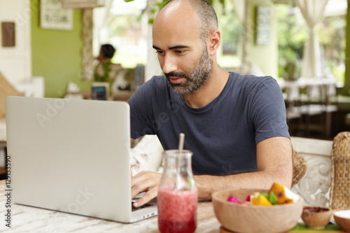 Bearded middle-aged self-employed man sitting at cafe in front of generic laptop and looking at screen with serious and concentrated expression while working remotely on his project  using free wi-fi