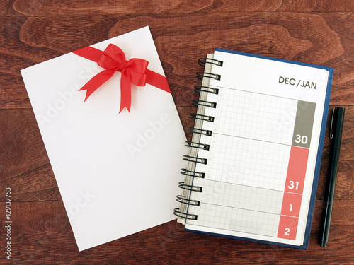 open calendar diary planner with date of December and January in grid and envelope with red ribbon bow and pen on desk floor, holiday event planning, flat lay close-up top view