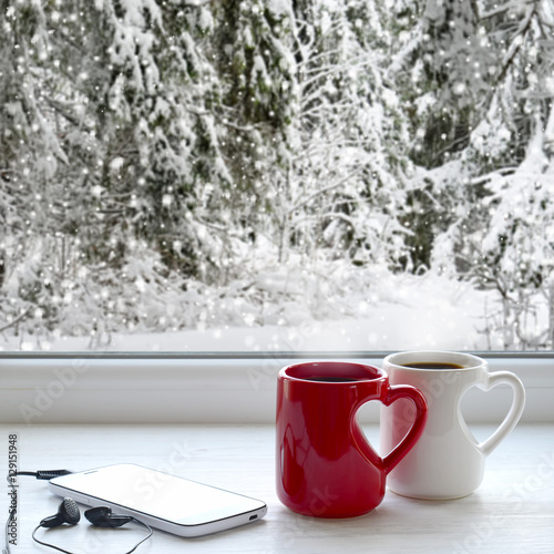 Two coffee cups, smartphone and headphones on a windowsill. In the background, a beautiful winter forest in snow