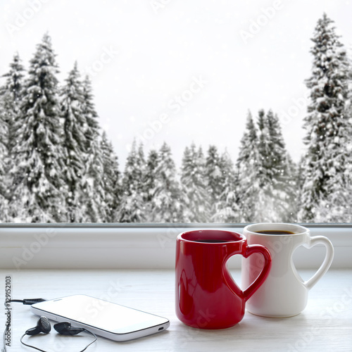 Two coffee cups, smartphone and headphones on a windowsill. In the background, a beautiful winter forest in snow