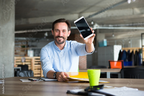 Portrait of happy smiling businessman showing mobile or smart phone with blank screen while working with documentation in office interior. Business ideas are welcomed.
