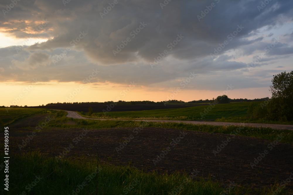 beautiful spring rural sunset over the field, separated by a sandy road, nature, landscape, bright sky and dark clouds 