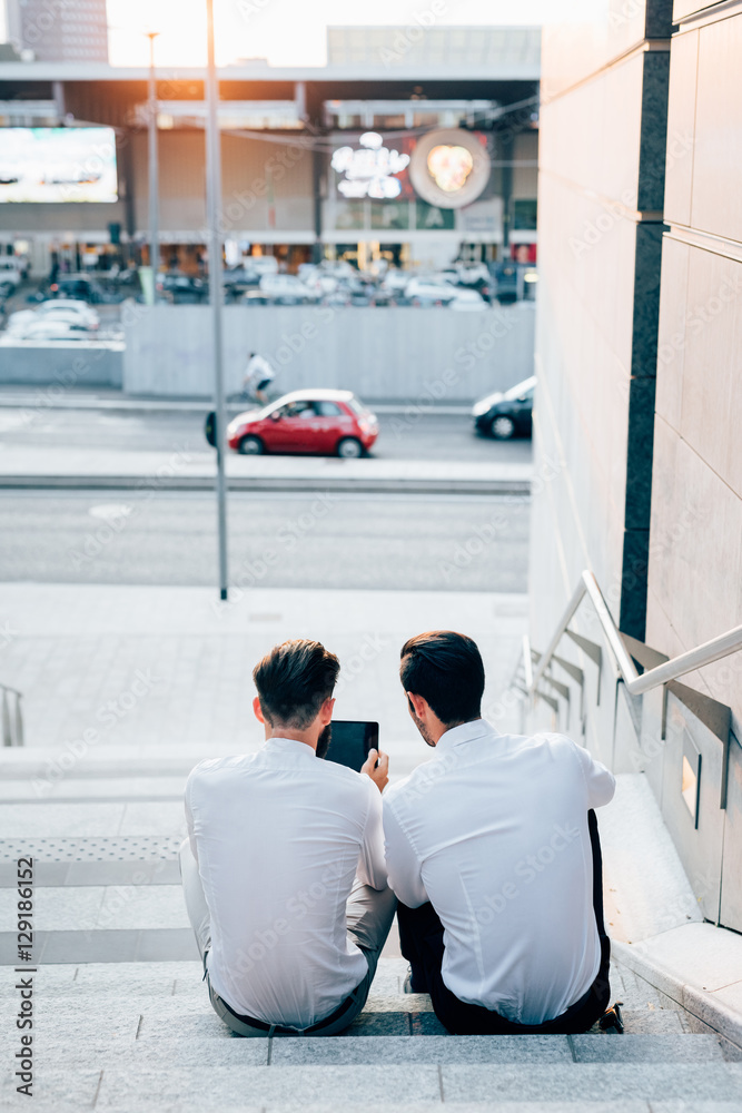 View from behind of two young bearded blonde and black hair modern businessman, sitting on a staircase,  using tablet - technology, business, work concept