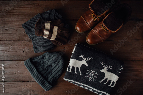 Male winter style on a brown wooden background. Winter brown shoes, grey sweater with deer, grey knitted scarf and hat and brown gloves. Top view