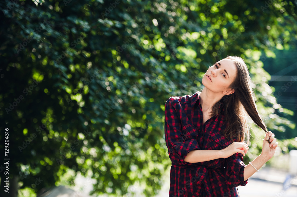 Cute teenage girl in casual dress playing with her braids, looking at camera and smiling, standing against green foliage Cute teenage girl in casual dress playing with her braids, looking at camera and smiling, standing against green foliage