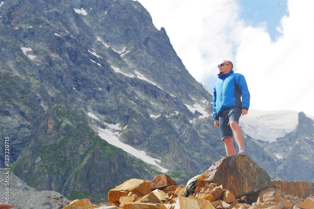 Young tourist in sunglasses, shorts, blue jacket stands on edge of cliff, admiring mountain scenery on background of snow-capped peaks, floating sky clouds. Sport and concept of healthy lifestyle Young tourist in sunglasses, shorts, blue jacket stands on edge of cliff, admiring mountain scenery on background of snow-capped peaks, floating sky clouds. Sport and concept of healthy lifestyle