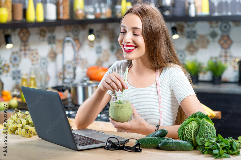 Young woman sitting on the kitchen table with laptop, smoothie and green vegetables. Healthy eating, weight loss and detox concept Young woman sitting on the kitchen table with laptop, smoothie and green vegetables. Healthy eating, weight loss and detox concept
