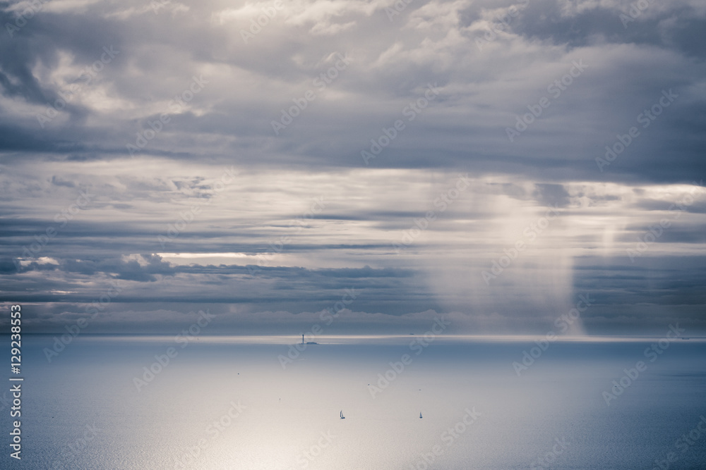 la mer bleue sous un ciel bleu et blanc avec un phare, des bateaux et la pluie au loin