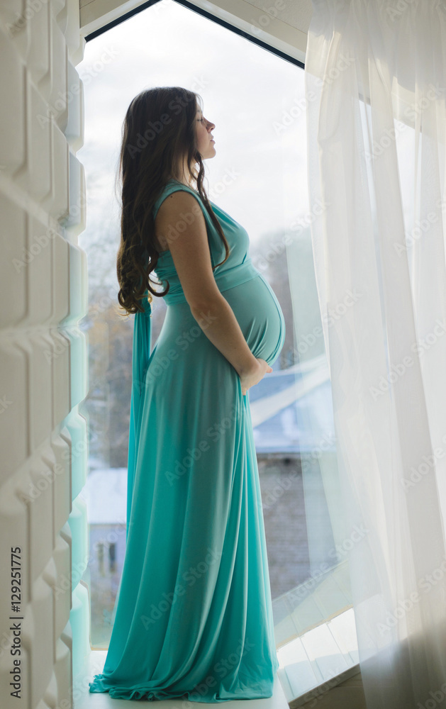 Pregnant girl standing against wall in azure dress near window Pregnant girl standing against wall in azure dress near window