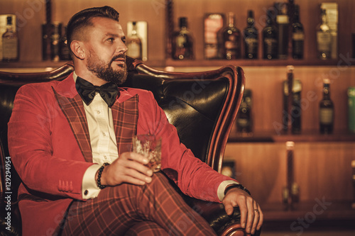 Extravagant stylish man with whisky glass sitting on armchair in
