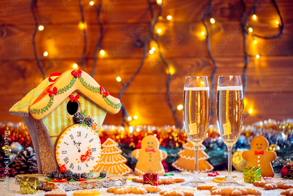 Christmas Gingerbread House with clock and two glass champagne with fir festive decoration with snow on blurred bokeh background.