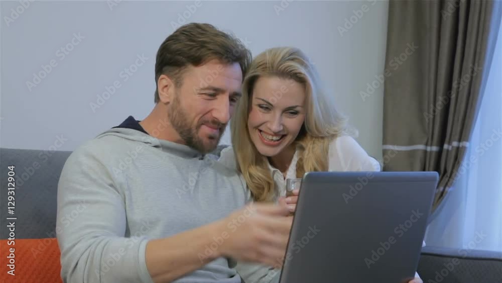 Nice caucasian couple looking at laptop at home. Attractive bearded man showing something on laptop to his pretty blond wife. Handsome brunette man pointing his hand on the laptop screen Nice caucasian couple looking at laptop at home. Attractive bearded man showing something on laptop to his pretty blond wife. Handsome brunette man pointing his hand on the laptop screen