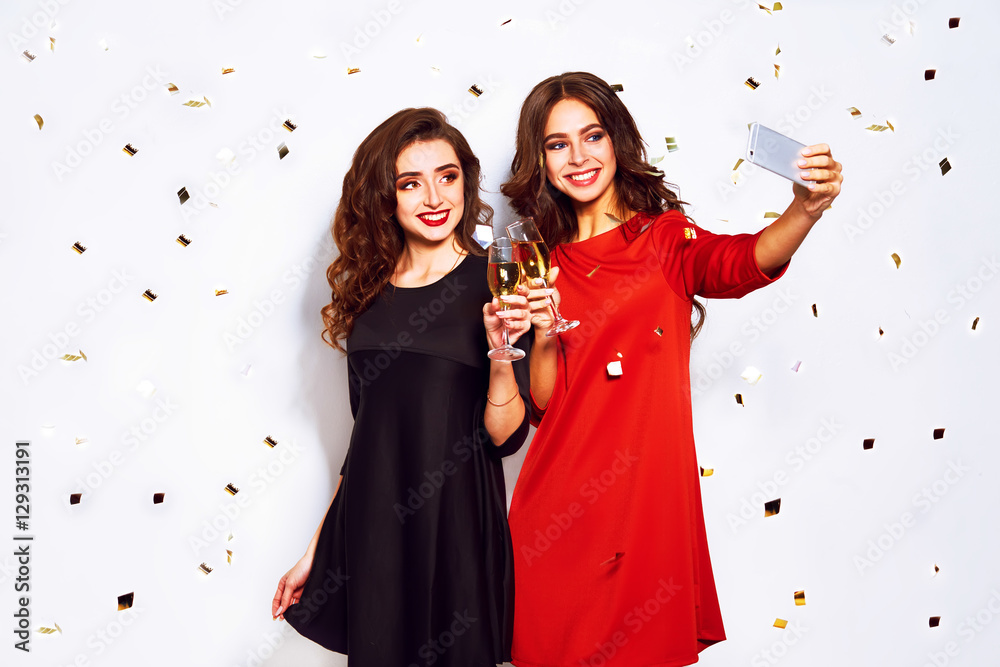 Plakat portrait of two happy young women who make selfie. in evening dresses on party over white background. firecrackers in the background.confetti.the concept party.funny faces
