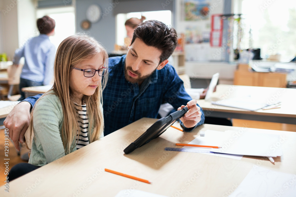 Teacher discussing with student while showing digital tablet at desk in classroom Teacher discussing with student while showing digital tablet at desk in classroom