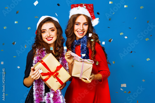 portrait of two happy young women in santa claus hat with gift .Christmas concept. in evening dresses on party over blue background. firecrackers in the background. 