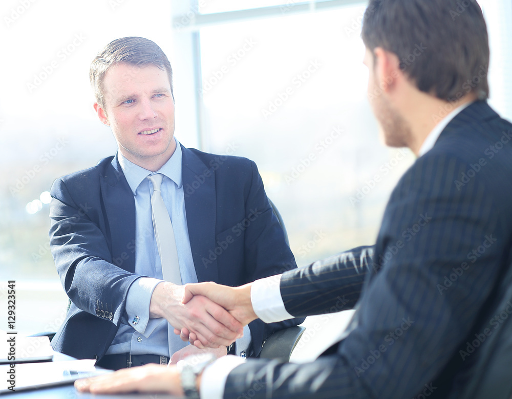 Business colleagues sitting at a table during a meeting with two male executives shaking hands Business colleagues sitting at a table during a meeting with two male executives shaking hands