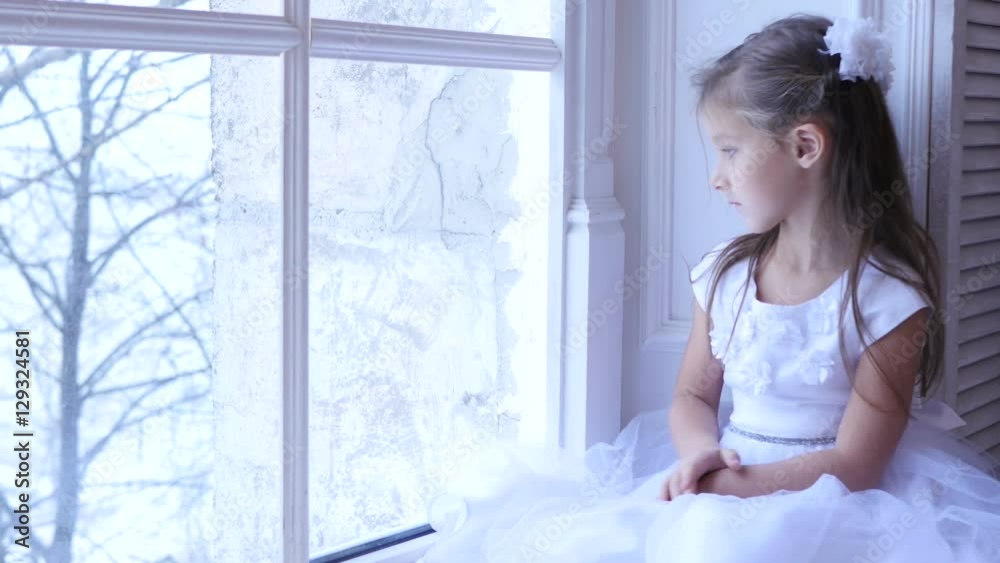 Sad little girl looking the window, kid looks at  snowing landscape