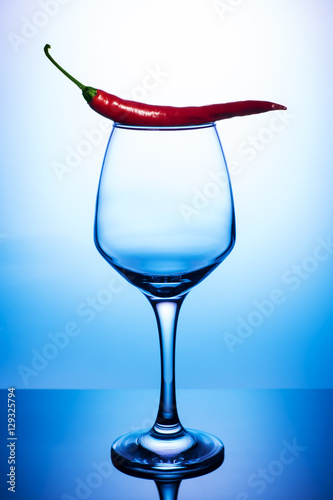 Empty wine glass with red hot chili pepper on a blue background. Transparent glass, vertical  photo.