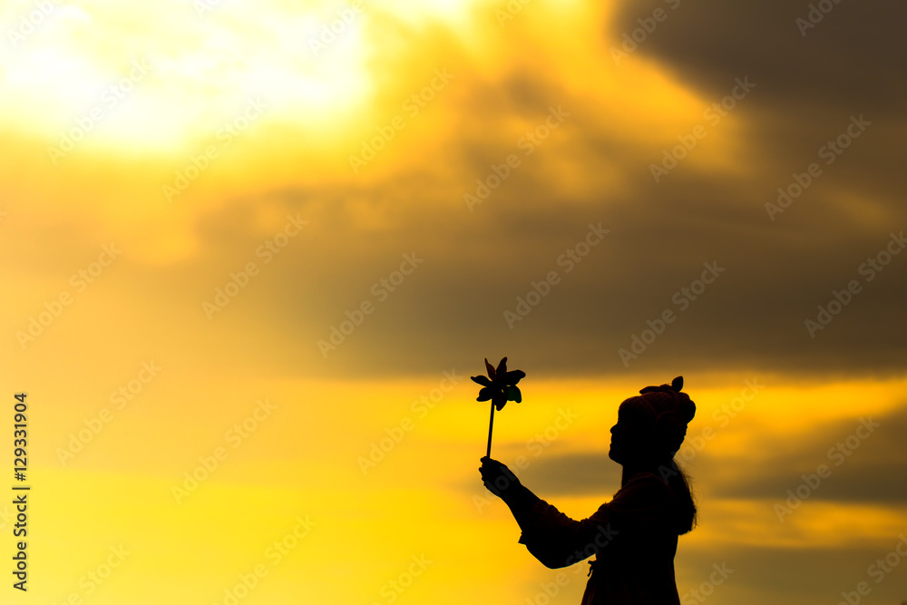 Silhouette of Beautiful girl holding wind toy or wind turbine or pinwheel and wool hat with golden sky on winter season in morning.