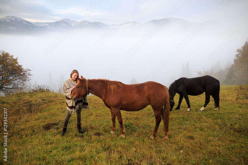 Beautiful girl with horses at mountain peak above clouds and fog. Girl with an horse playing together at the farm. Young woman take care of her horse. Early morning.Misty mountain.