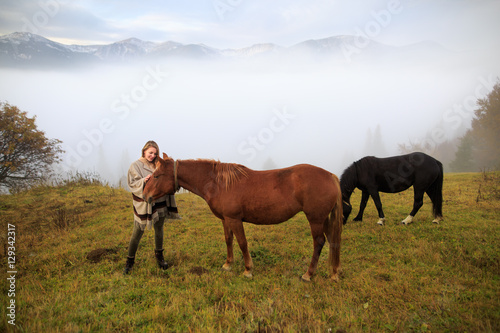 Beautiful girl with horses at mountain peak above clouds and fog. Girl with an horse playing together at the farm. Young woman take care of her horse. Early morning.Misty mountain.