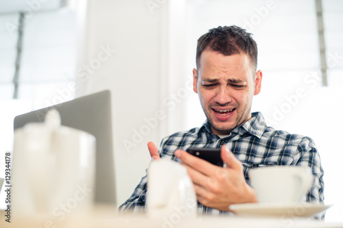 Bad news. Angry young man holding a mobile phone and grimacing at cafe