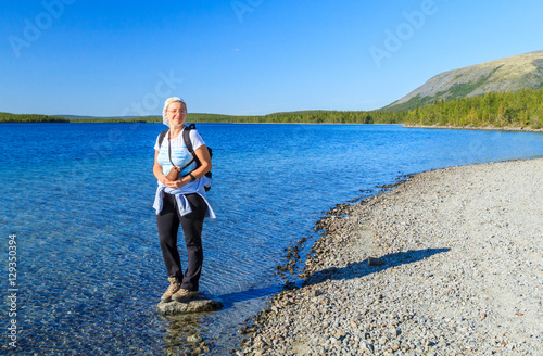 Woman traveler in outdoor clothes stands on a rock on the shore of a mountain lake above the Arctic Circle.