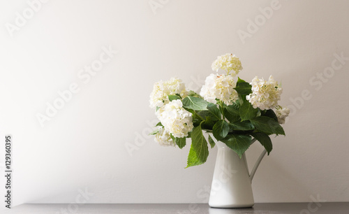 White hydrangeas in jug on black table against white wall (selective focus)