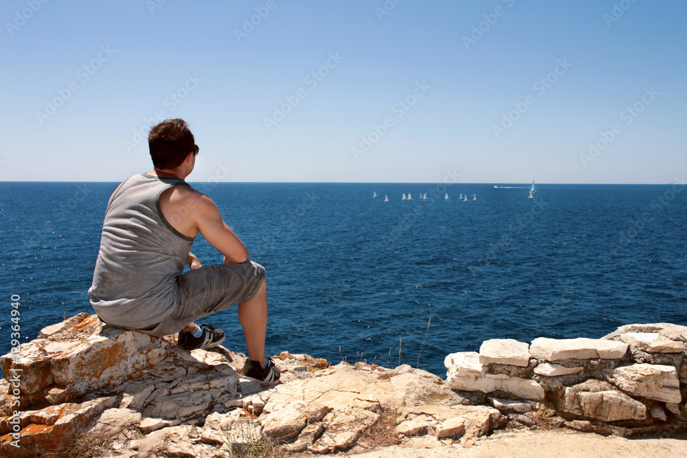 He is sitting and watching the boats sailing on the sea